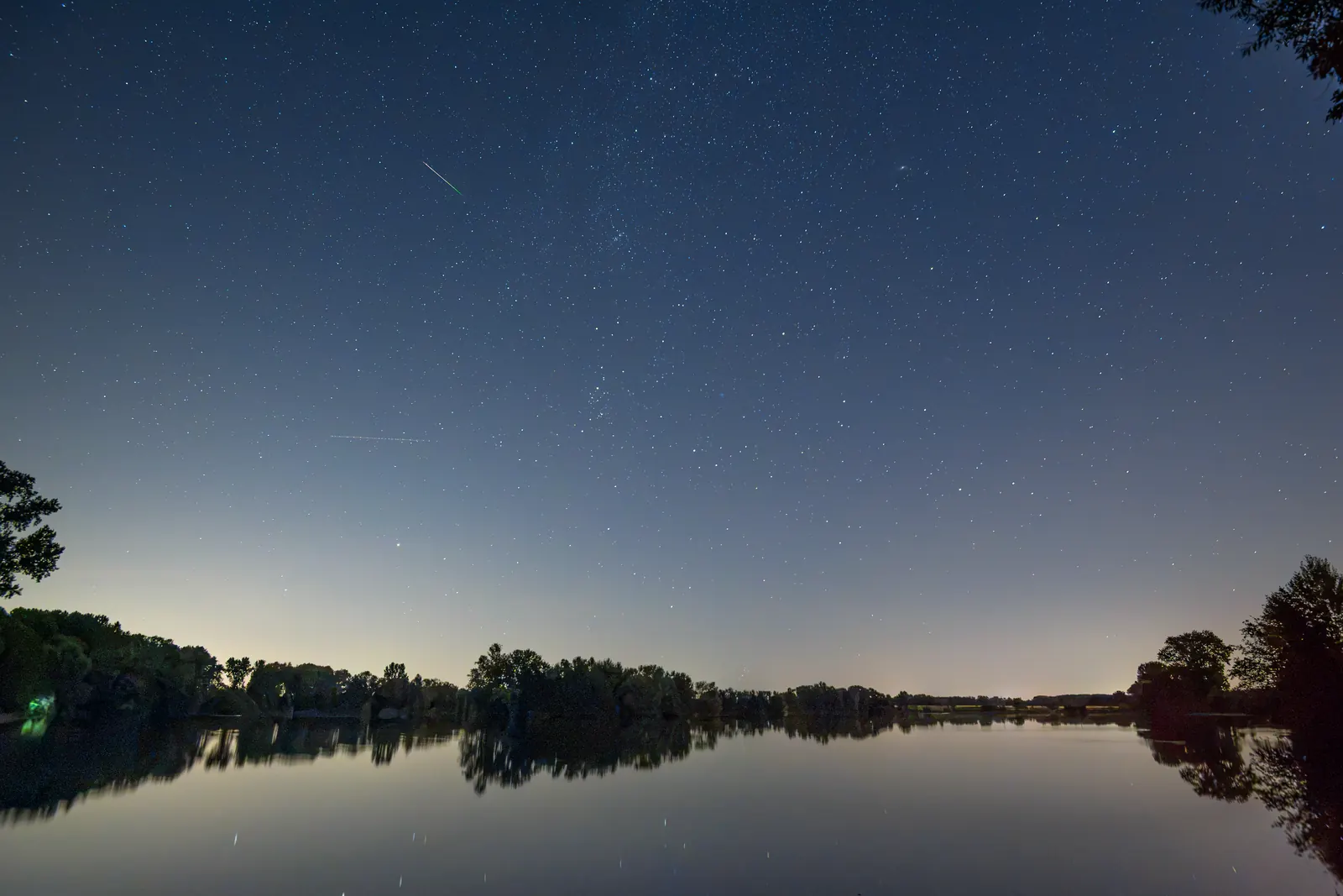 Perseiden-Meteor als feiner Strich über einem stillen See, Ufer im Spiegelbild
