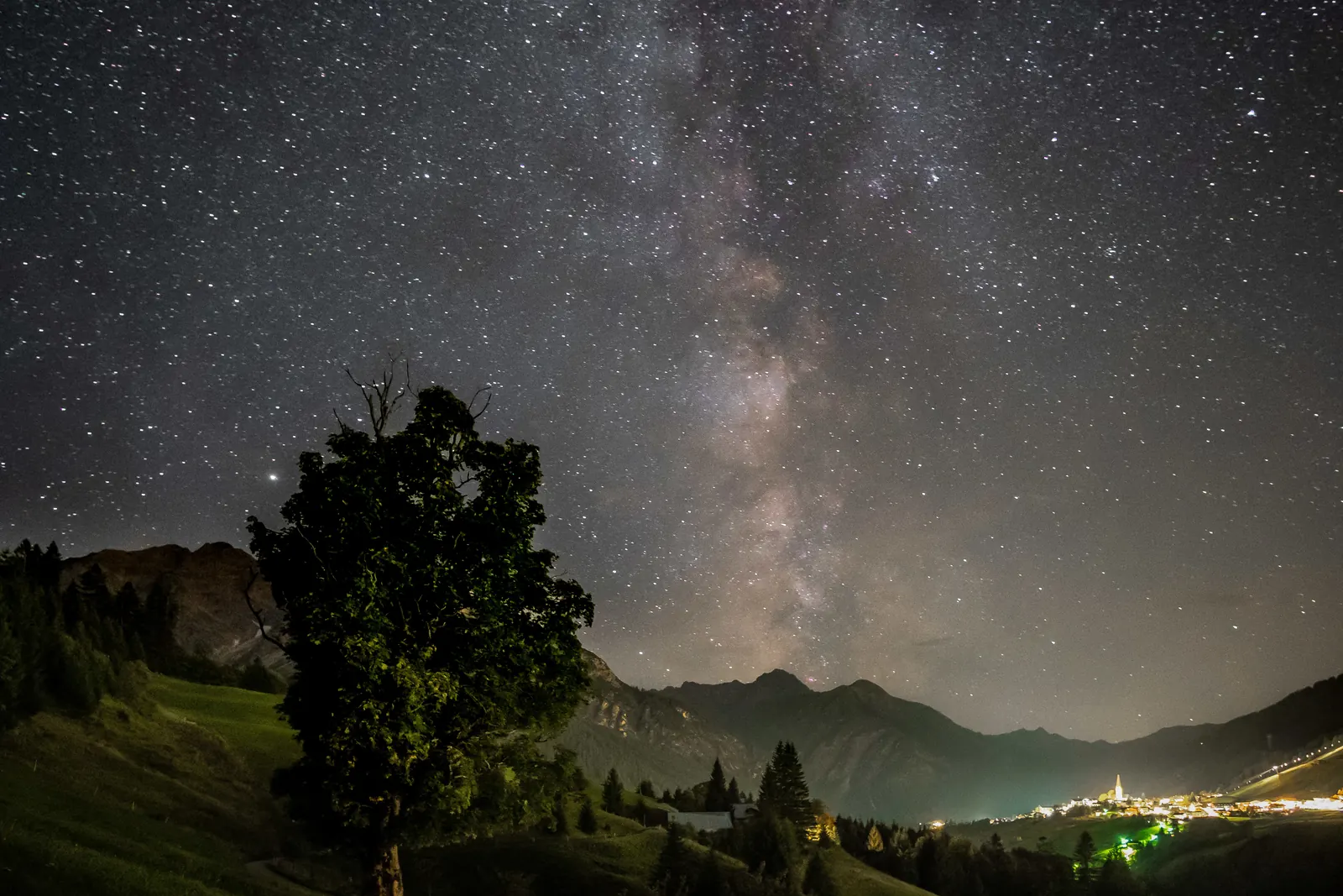 Milchstraße über einem Baum und Bergen im Kleinwalsertal, unten die Lichter eines Ortes